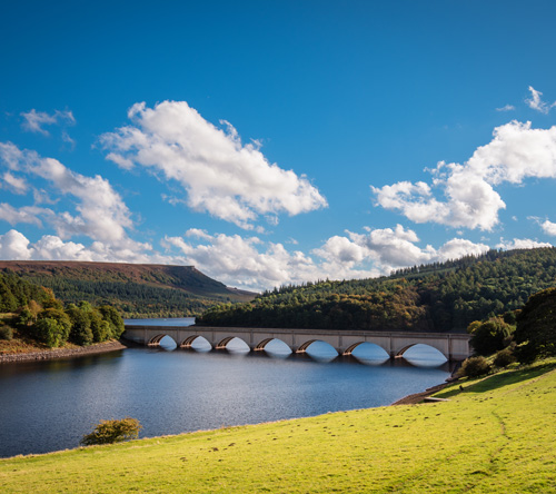 scenic view of a bridge spanning a lake surrounded by green hills and blue skies with clouds showcasing natural beauty and outdoor recreation